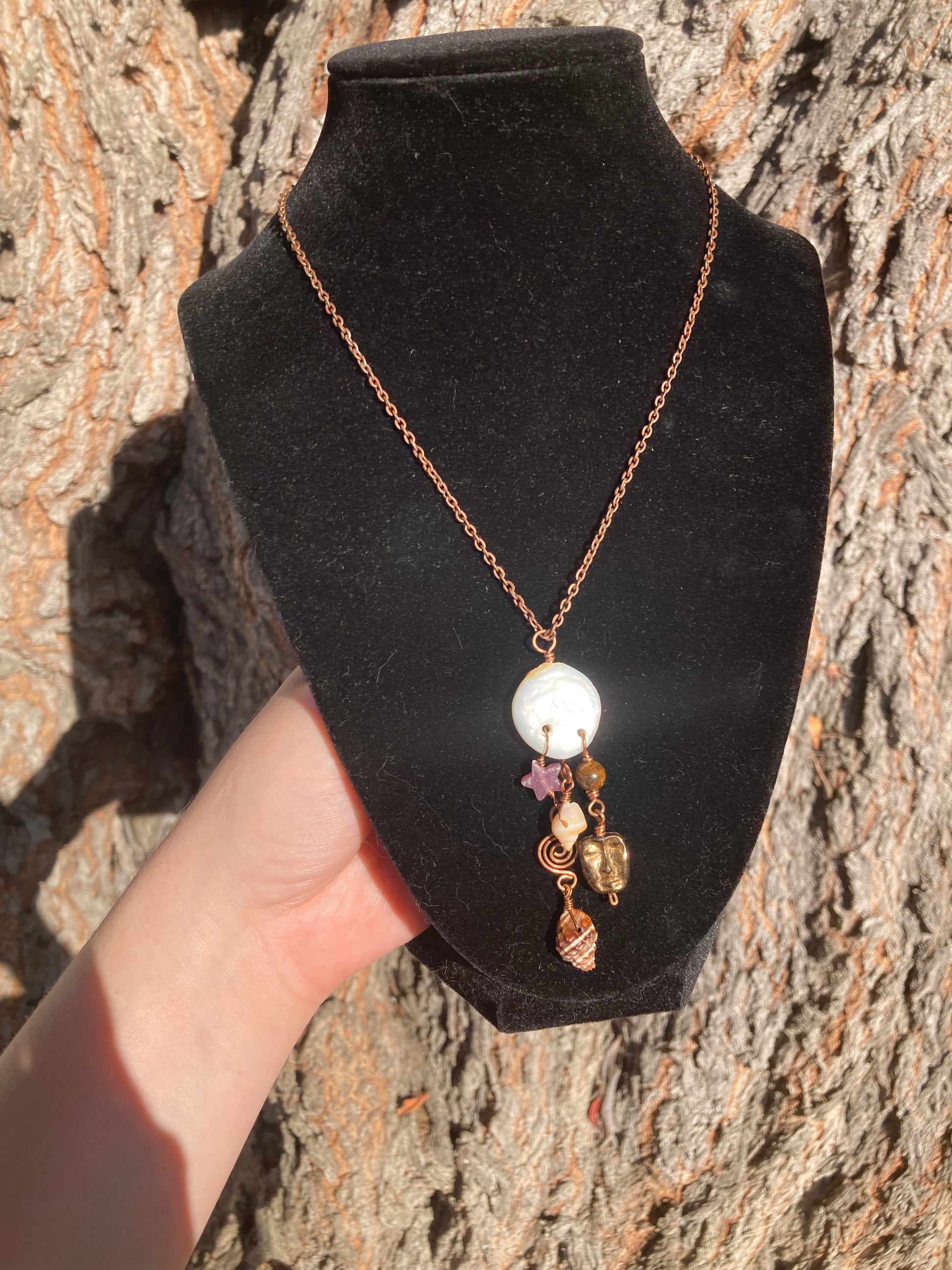 A copper chain necklace with a white shell pendant and small dangling charms, displayed on a black velvet jewelry bust held by a hand against a textured tree bark background.