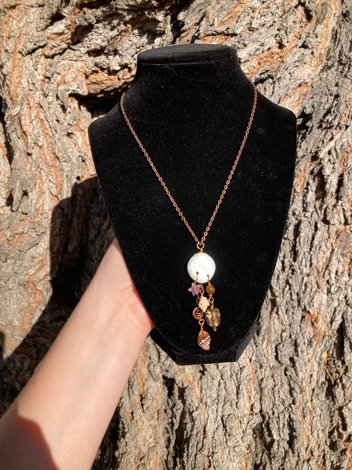 A copper chain necklace with a white round bead and dangling amber and purple crystal charms, displayed on a black velvet jewelry bust held against a textured tree trunk background.