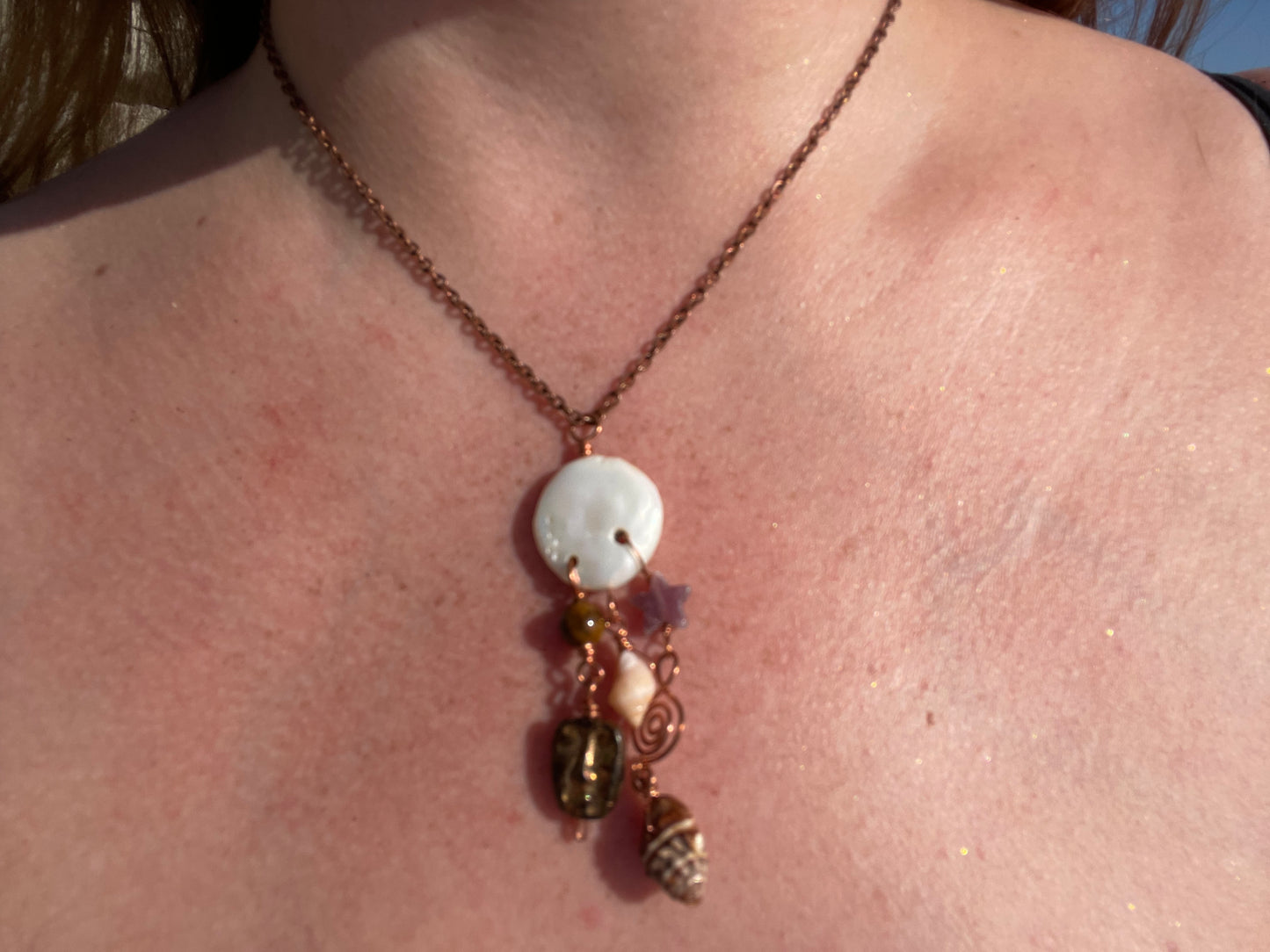 A close-up of a person's chest wearing a copper-toned chain necklace with a white shell pendant and dangling charms including a small star and acorn.