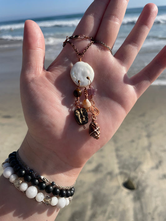 A handmade bohemian-style necklace with a white shell pendant, seashell charms, and copper wire details, held in a person's hand on a sandy beach with the ocean in the background.