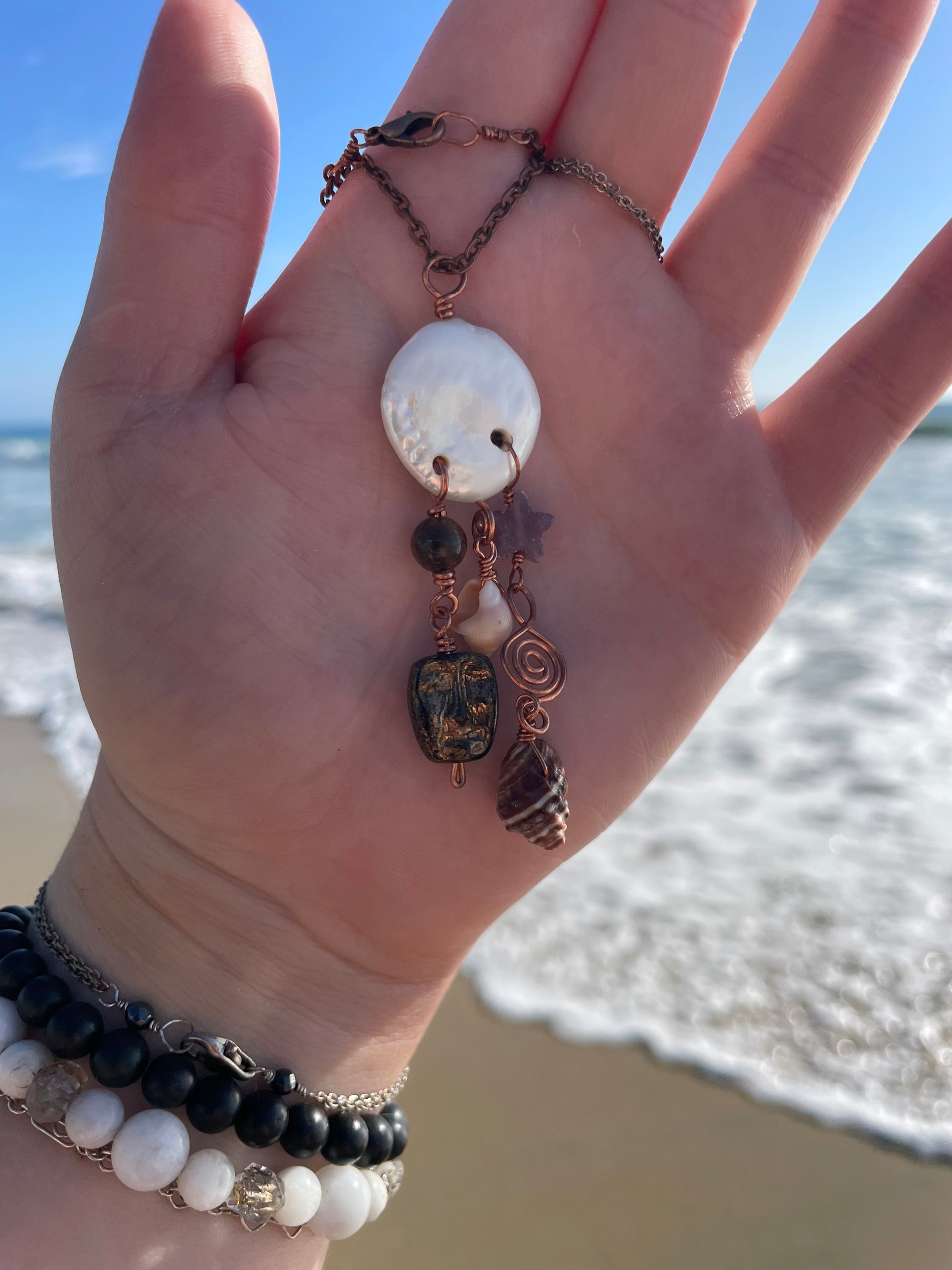 A handmade bohemian-style necklace with a white mother-of-pearl shell pendant, copper wire, and dark beads, held in a person's hand on a beach with ocean waves in the background.