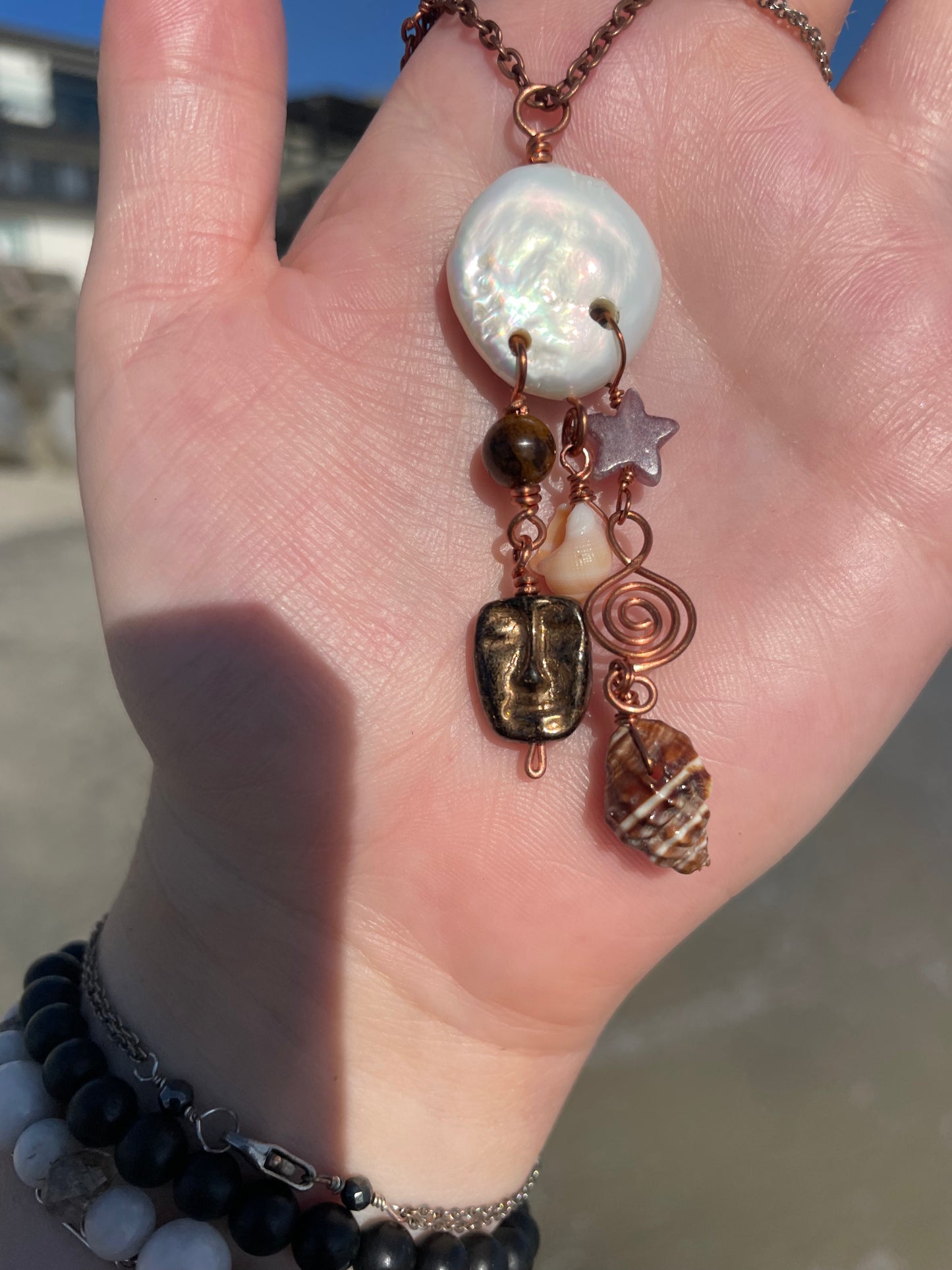 A close-up of a hand holding a bohemian-style necklace with a white mother-of-pearl disc, copper-toned beads, a bronze face pendant, a spiral wire charm, and a shell charm, against an outdoor beach background.